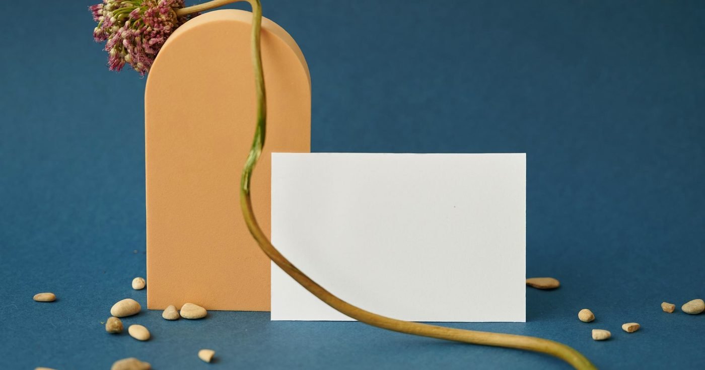 Tarjeta en blanco junto a un jarrón color durazno y una flor sobre un fondo azul intenso